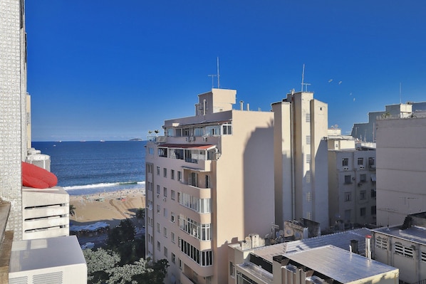 A high-rise building with balconies overlooking a beach, clear blue sky, and ocean view.