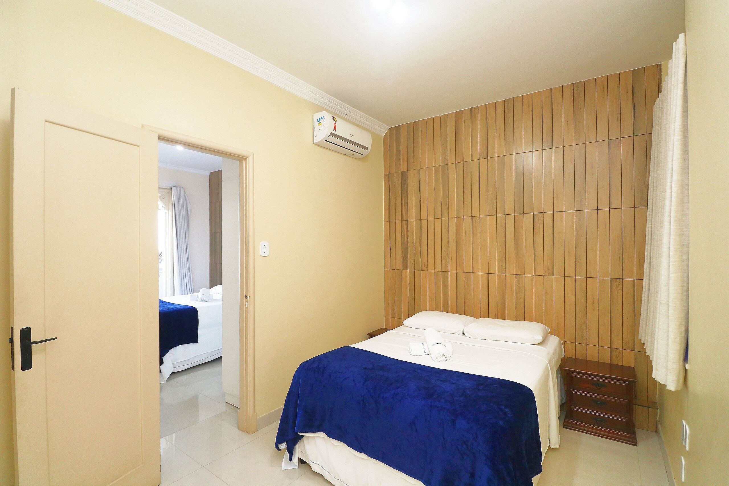 A hotel room with a bed, a wooden panel wall, an air conditioning unit, and a door leading to another room.