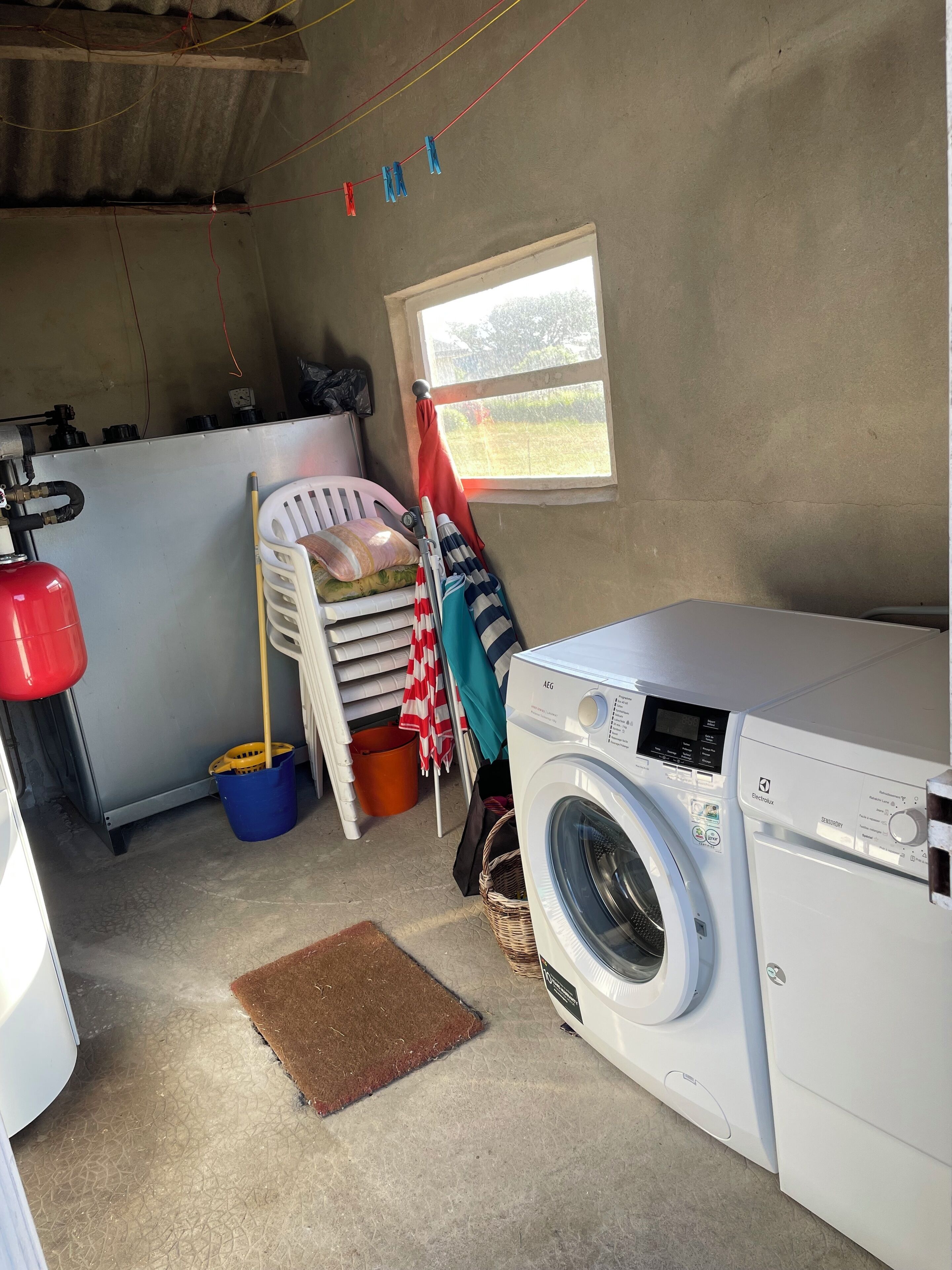 A laundry room with a washing machine, a drying rack, a bucket, and a window with a view of the outside.
