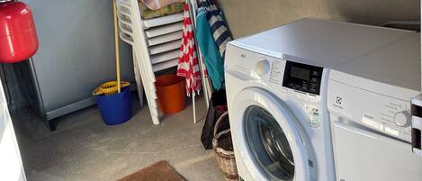 A laundry room with a washing machine, a drying rack, a bucket, and a window with a view of the outside.