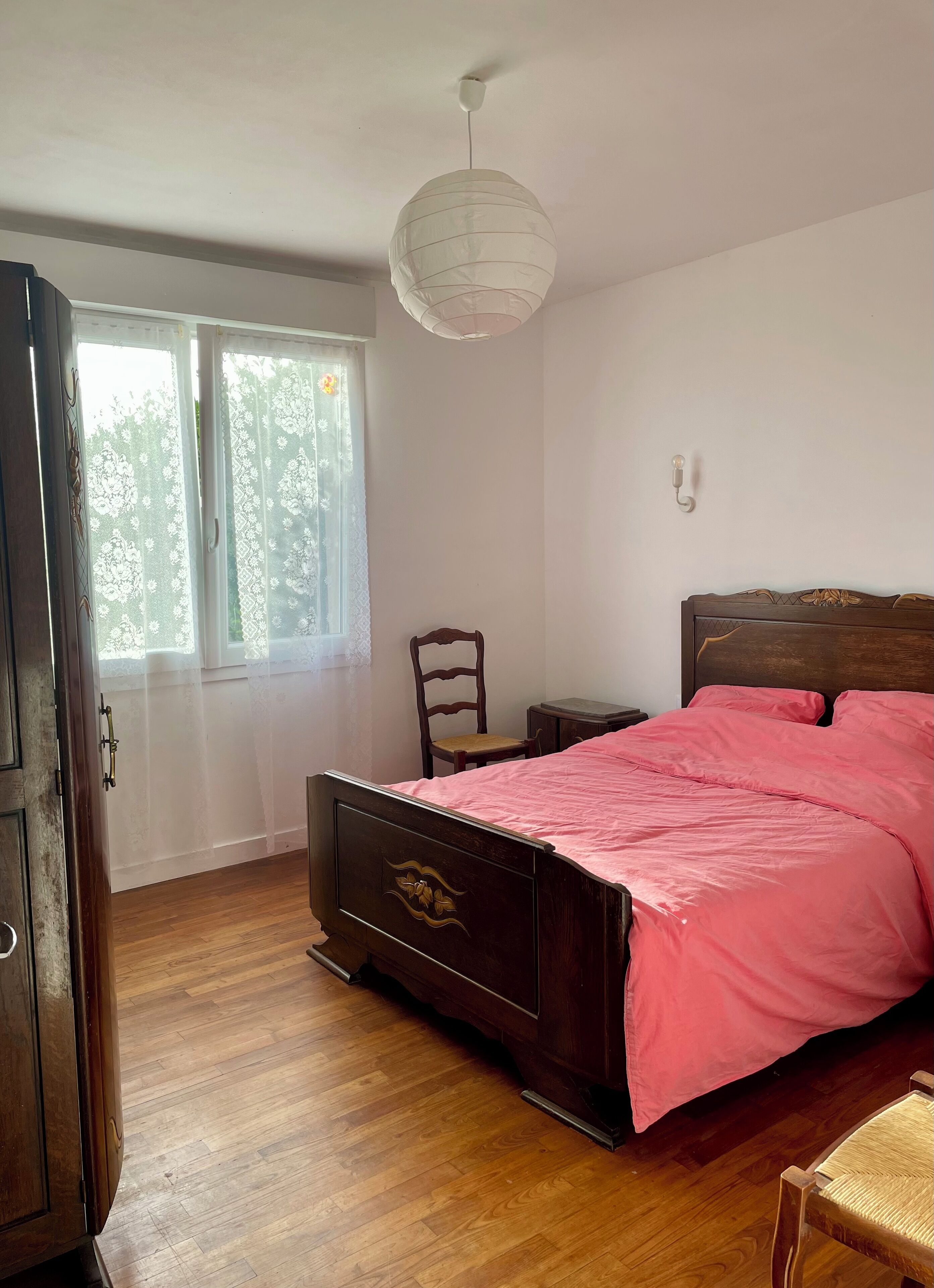 Une chambre à coucher avec un lit en bois, une chaise, une petite table et une fenêtre ornée de rideaux en dentelle.