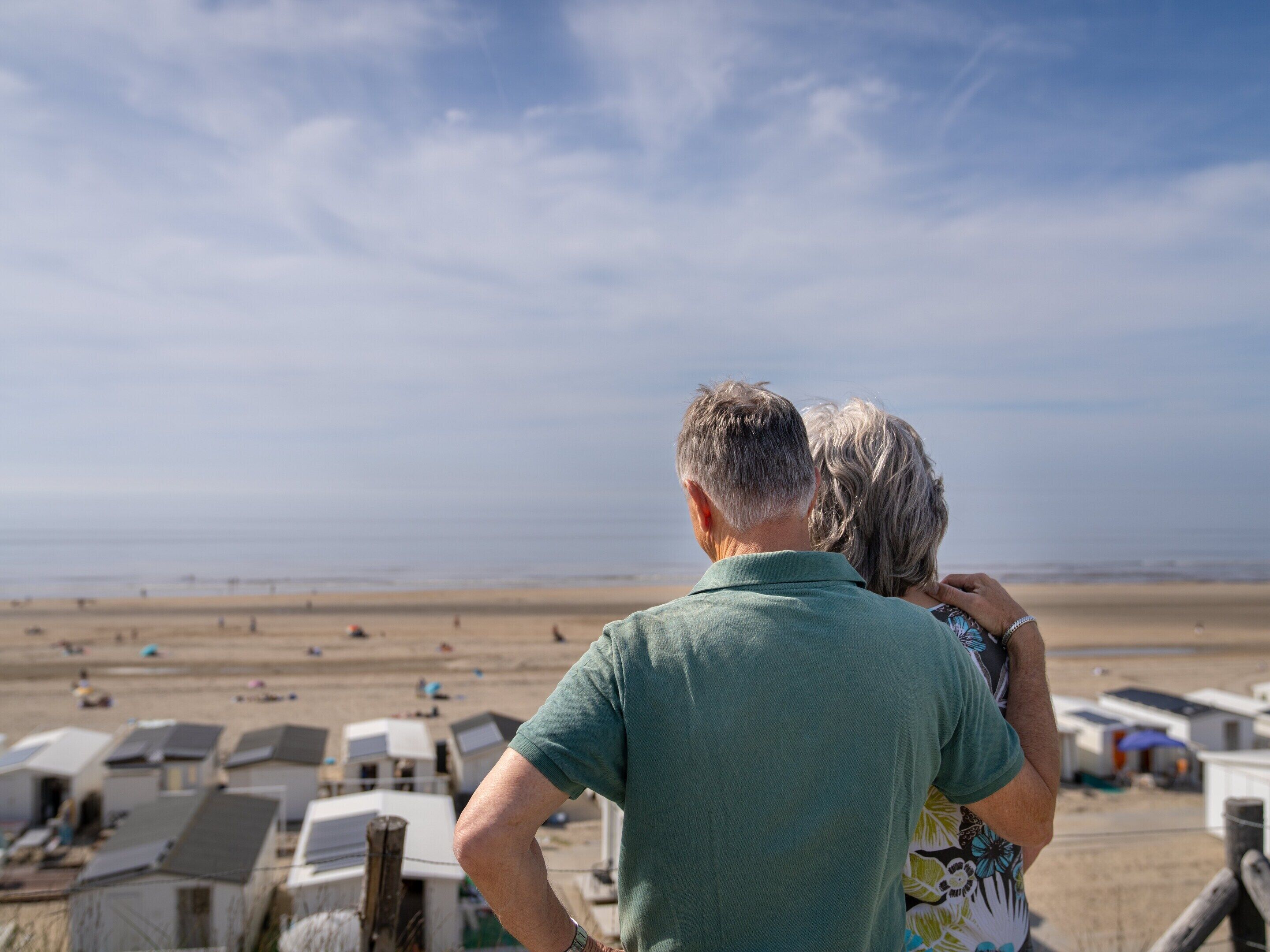 Un couple se tient dos à l’appareil photo, surplombant une plage où l’on aperçoit plusieurs camping-cars et tentes au loin.