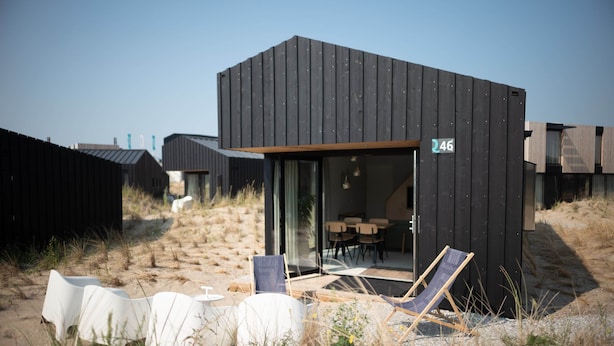 A modern beach house with a black exterior, numbered 46, featuring a glass door entrance, outdoor seating with white chairs, and a wooden deck.