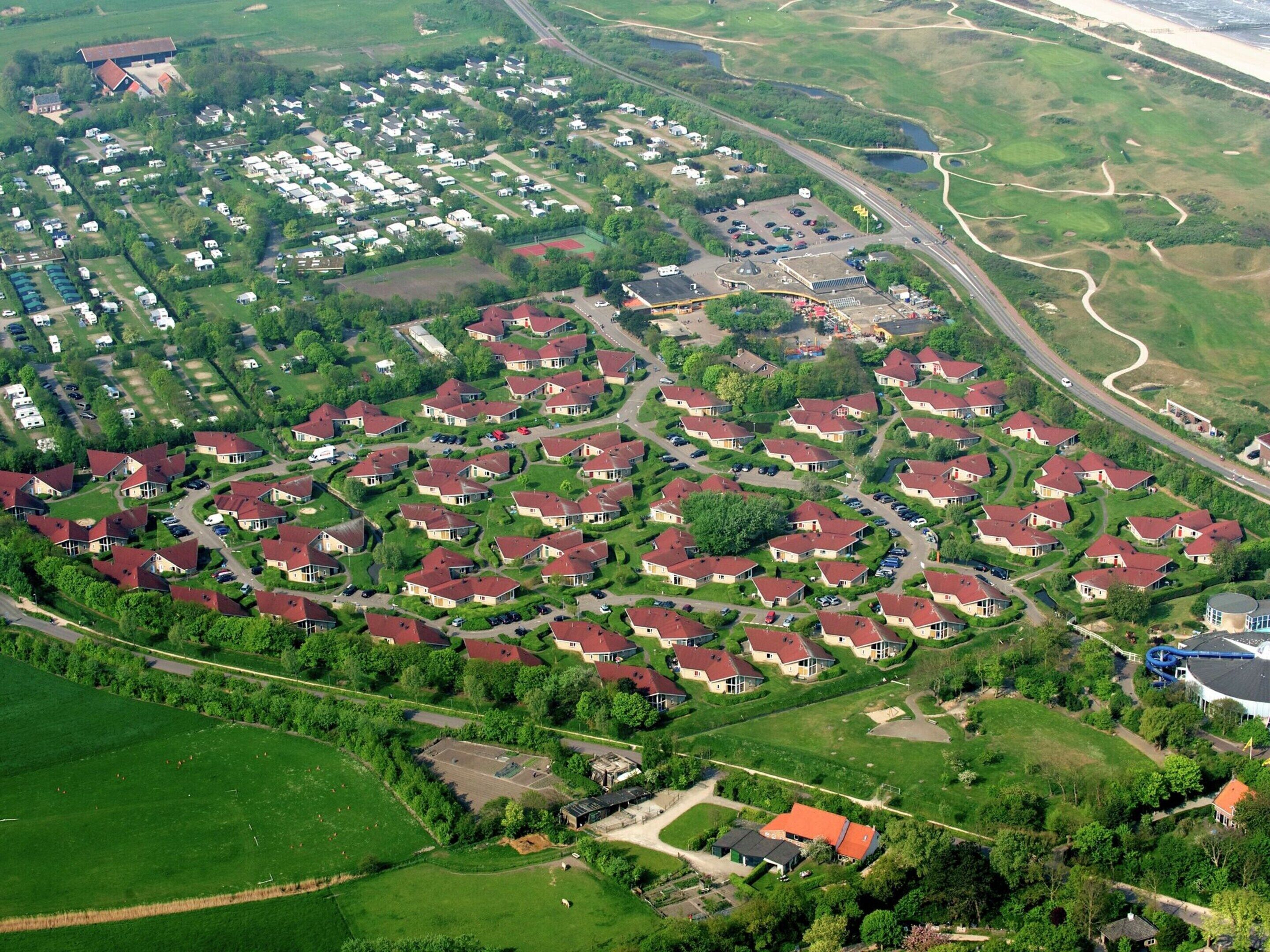 An aerial view of a residential area with houses arranged in a grid pattern, surrounded by green fields and a road running through the center.