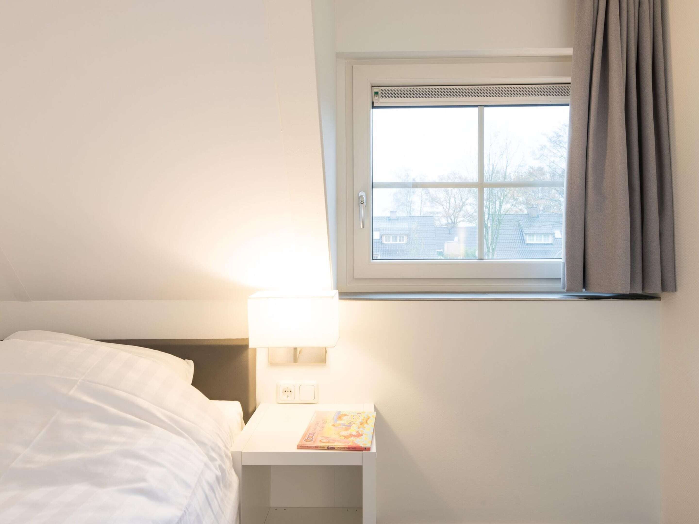 Una camera d'albergo con un letto, un comodino con una lampada, una finestra con tende e vista sugli edifici.