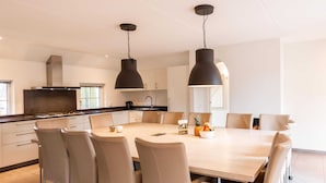 Une cuisine moderne dotée d’un îlot central, de luminaires suspendus noirs et d’un coin repas avec une table et des chaises aux couleurs claires.