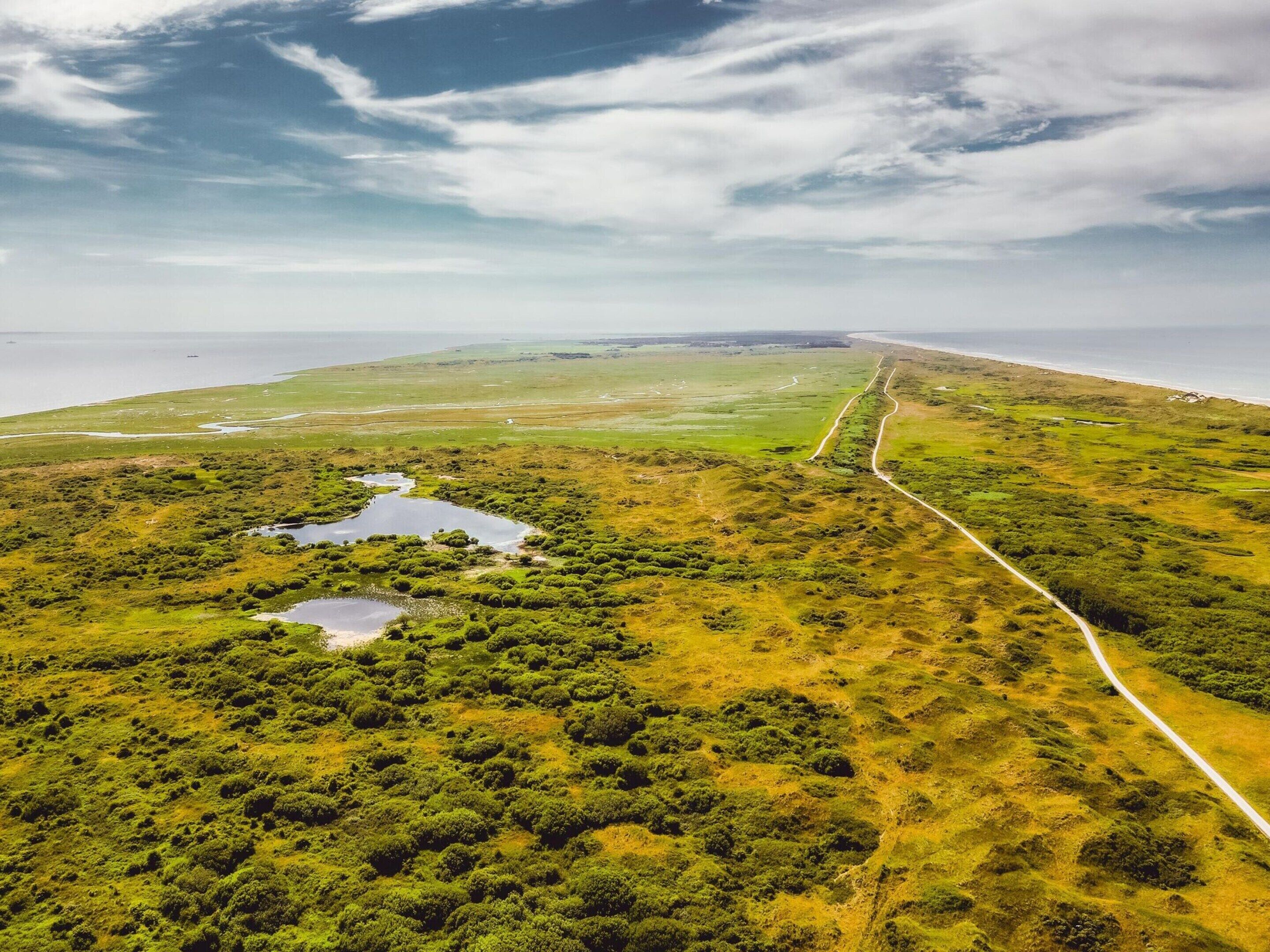 Luchtbeeld van een kustlandschap met een weg die naar het strand leidt, omgeven door groene begroeiing en kleine waterlichamen.