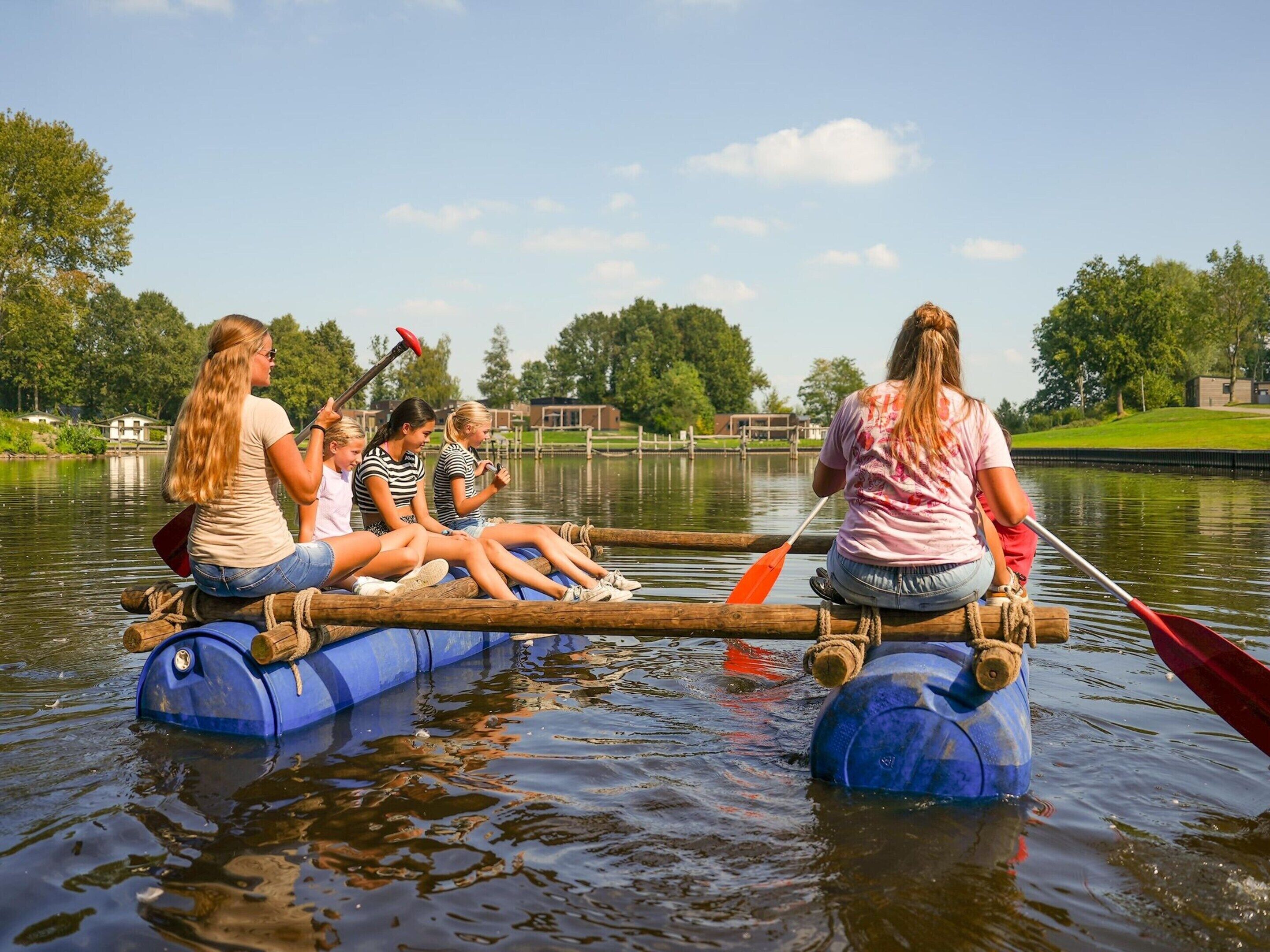 A group of people on a log raft in a calm body of water, surrounded by greenery and a clear sky.