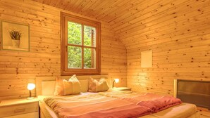 A wooden cabin bedroom with a bed, two bedside tables, a window, and a fireplace. - Holiday Home in Liebenfels in Carinthia (Liebenfels)
