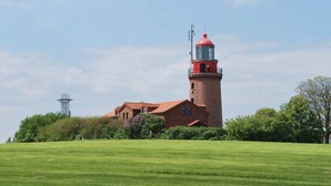 Un phare avec une base en briques et un sommet rouge, entouré d’un champ herbeux et d’arbres.