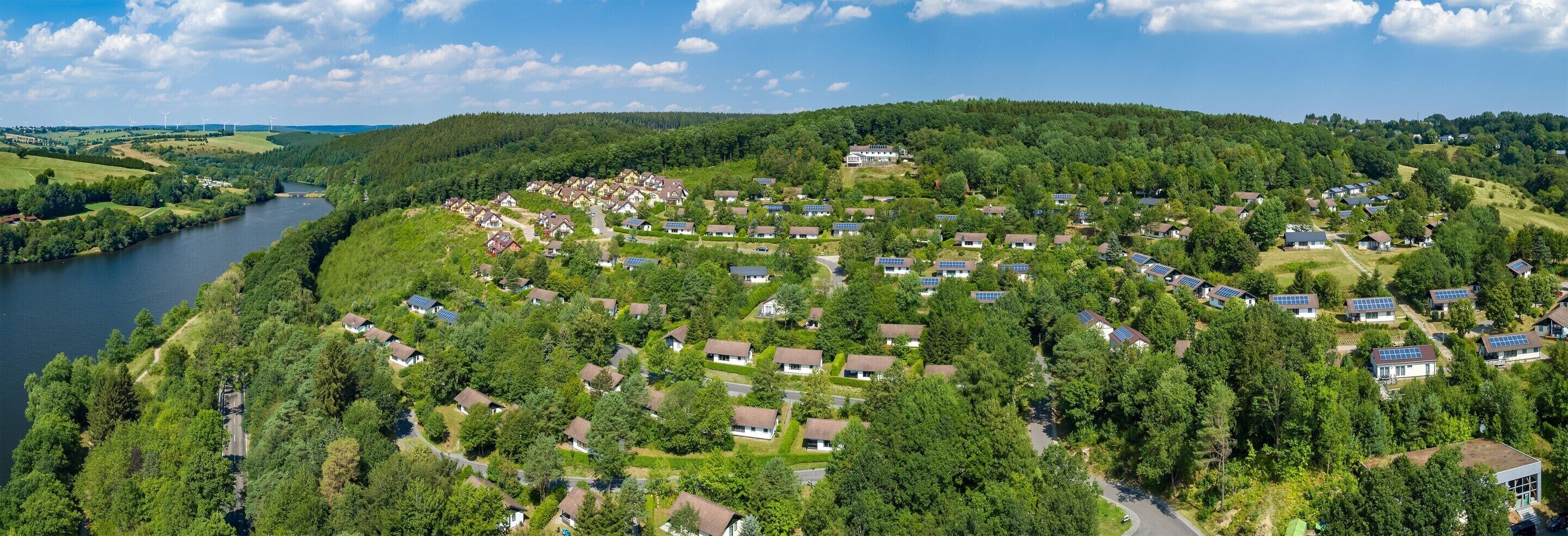 A residential area with houses surrounded by greenery, a river, and wind turbines in the distance.