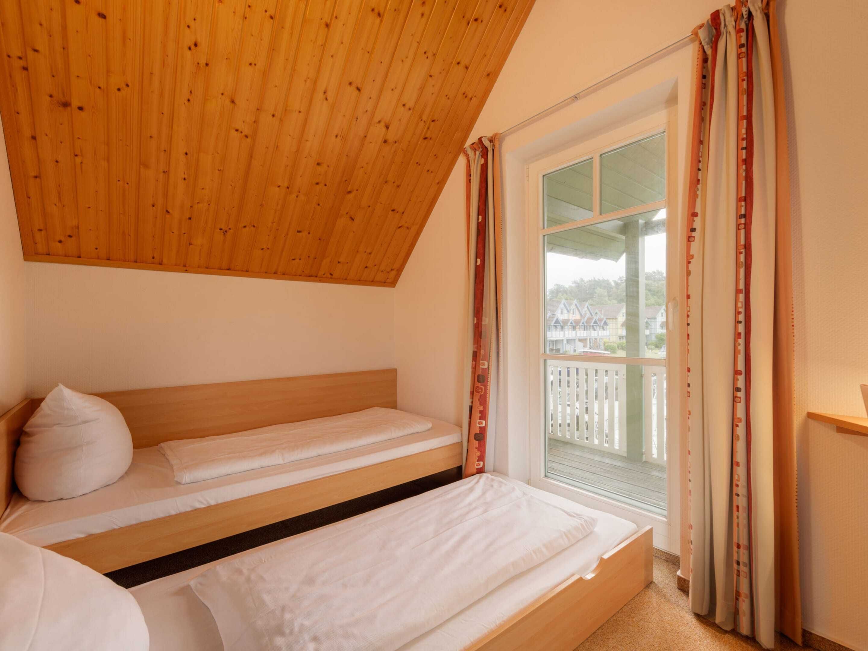 Ein Schlafzimmer mit schrägverdeckter Decke, einem großen Fenster mit Vorhängen und Blick auf Häuser.