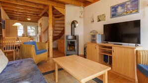 A cozy room with a wooden ceiling, a TV, a fireplace, wicker furniture, and a painting of a lighthouse. - Cozy Holiday Home with Wood Burning Stove near Water (Rheinsberg)
