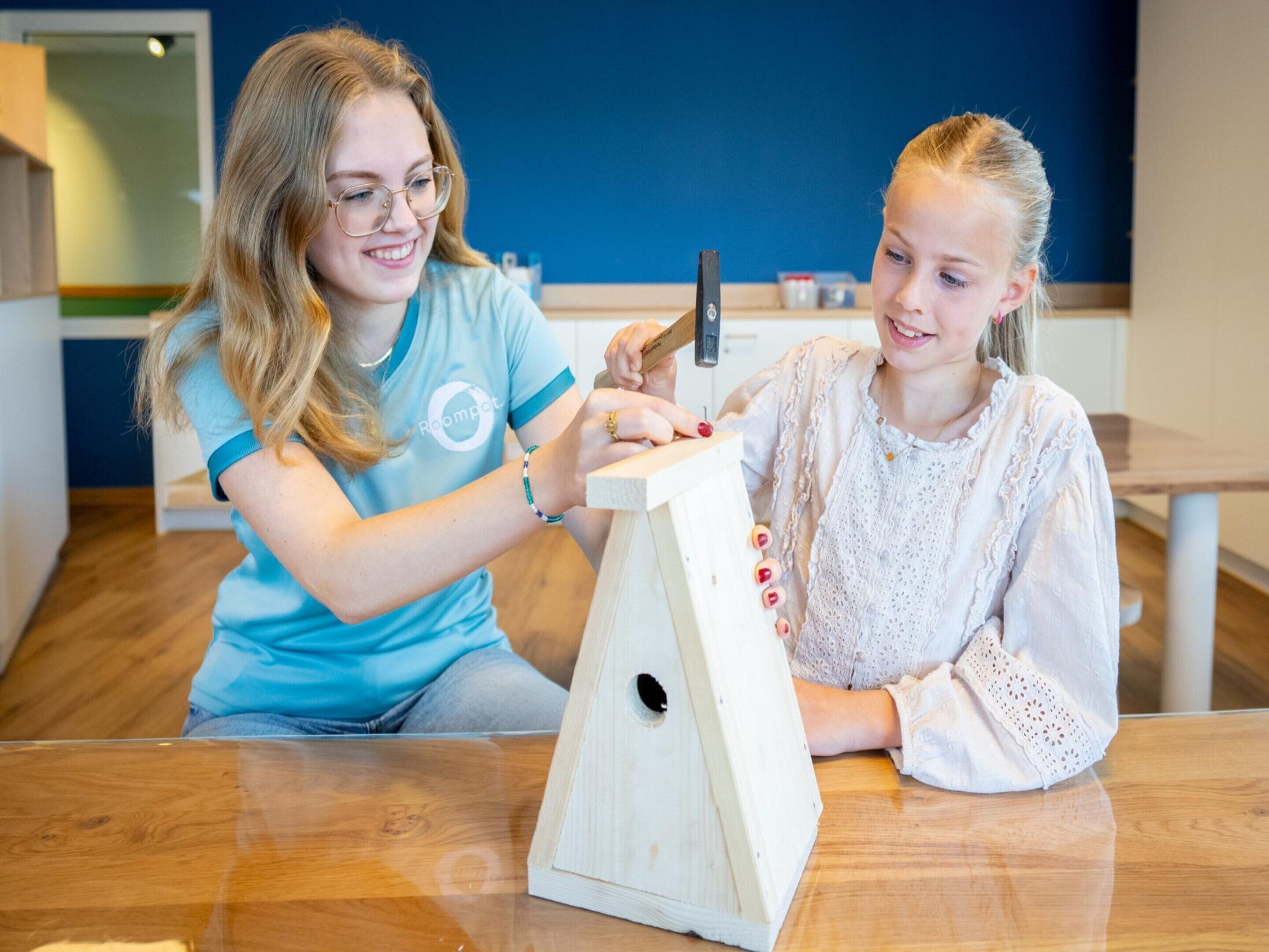 Two individuals engaged in a woodworking activity, seated at a wooden table with a simple wooden structure in front of them.