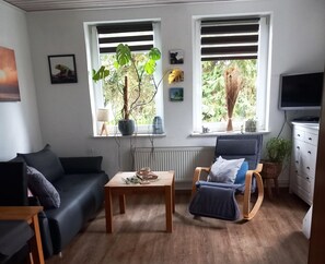 Un salon avec un canapé bleu foncé, une table basse en bois, un fauteuil à bascule et deux fenêtres équipées de stores.