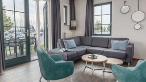 Een moderne woonkamer met een grijze hoekbank, teal fauteuils en een ronde salontafel. Grote ramen bieden uitzicht op een jachthaven met boten.
