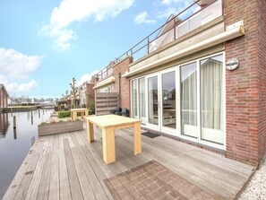 A wooden deck with a table and chairs, a brick building with large windows, and a calm water body in the background. - Nice Apartment with Dishwasher near Amsterdam (Uitgeest)