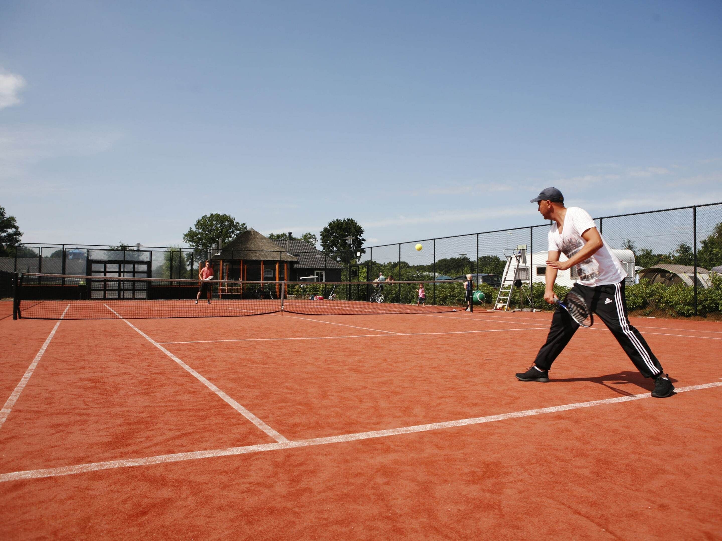 Ein Tennisplatz mit Netz, ein Spieler in weißem Hemd und schwarzer Hose sowie ein kleines Pavillon im Hintergrund.