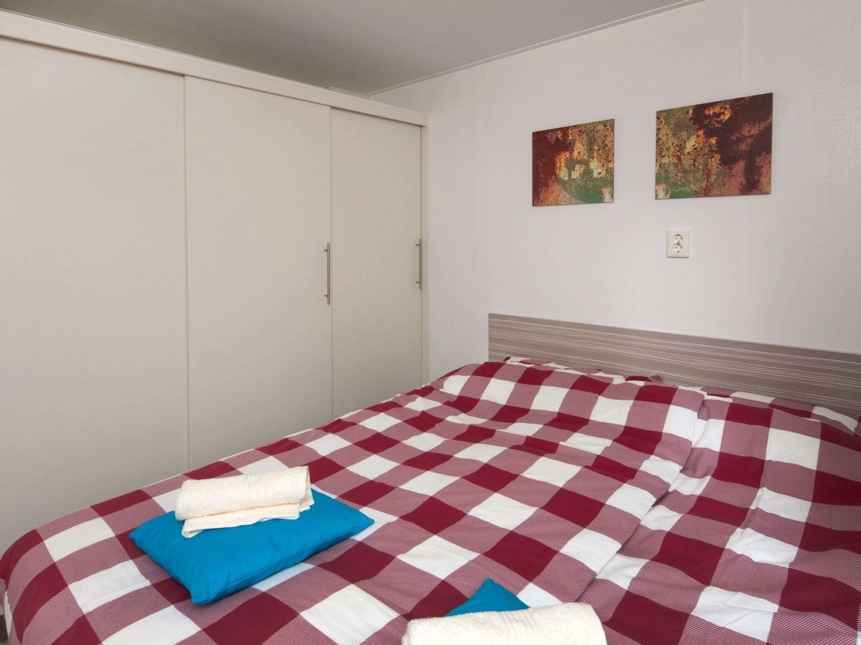 A bedroom with a checkered bedspread, a blue pillow, and a white towel. There are two paintings on the wall and a wardrobe with sliding doors.