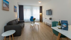 A modern living room with a grey sofa, a small white table, a blue chair, a TV mounted on the wall, and a dining area with a white table and chairs. - Apartment in Zoutelande Near the Beach (Zoutelande)