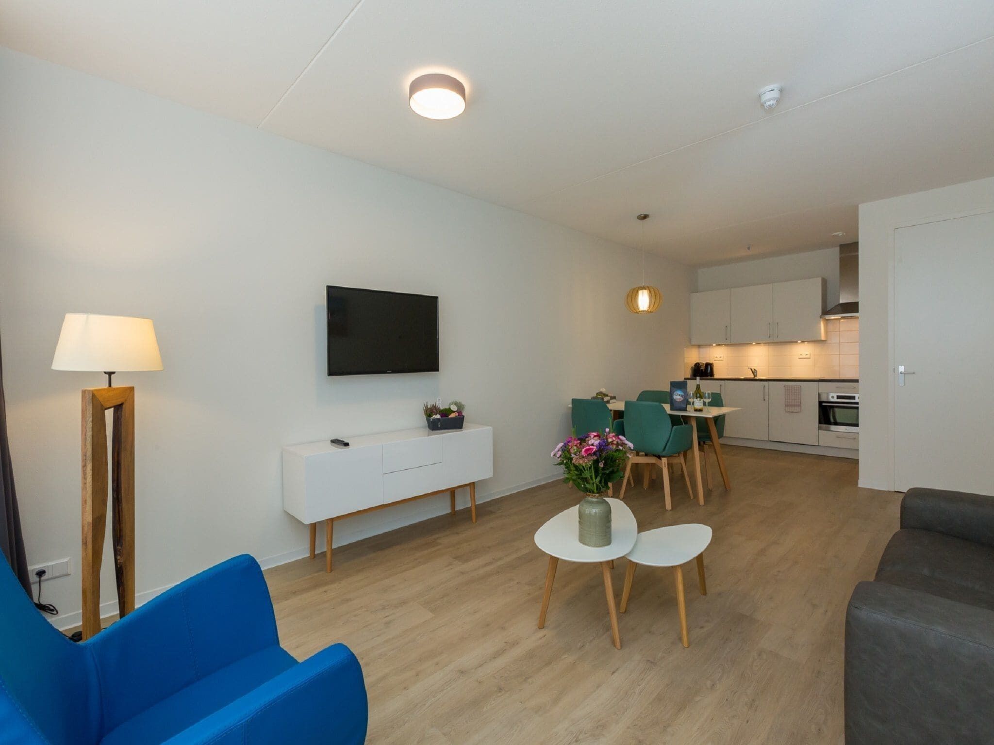 A modern living room with a flat-screen TV, a white console table, a blue armchair, and a small round table with a vase of flowers.