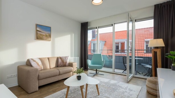A modern living room with a sofa, a small table, and a chair. There is a large window with a view of a red-roofed building outside. - Apartment in Zoutelande Near the Beach (Zoutelande)