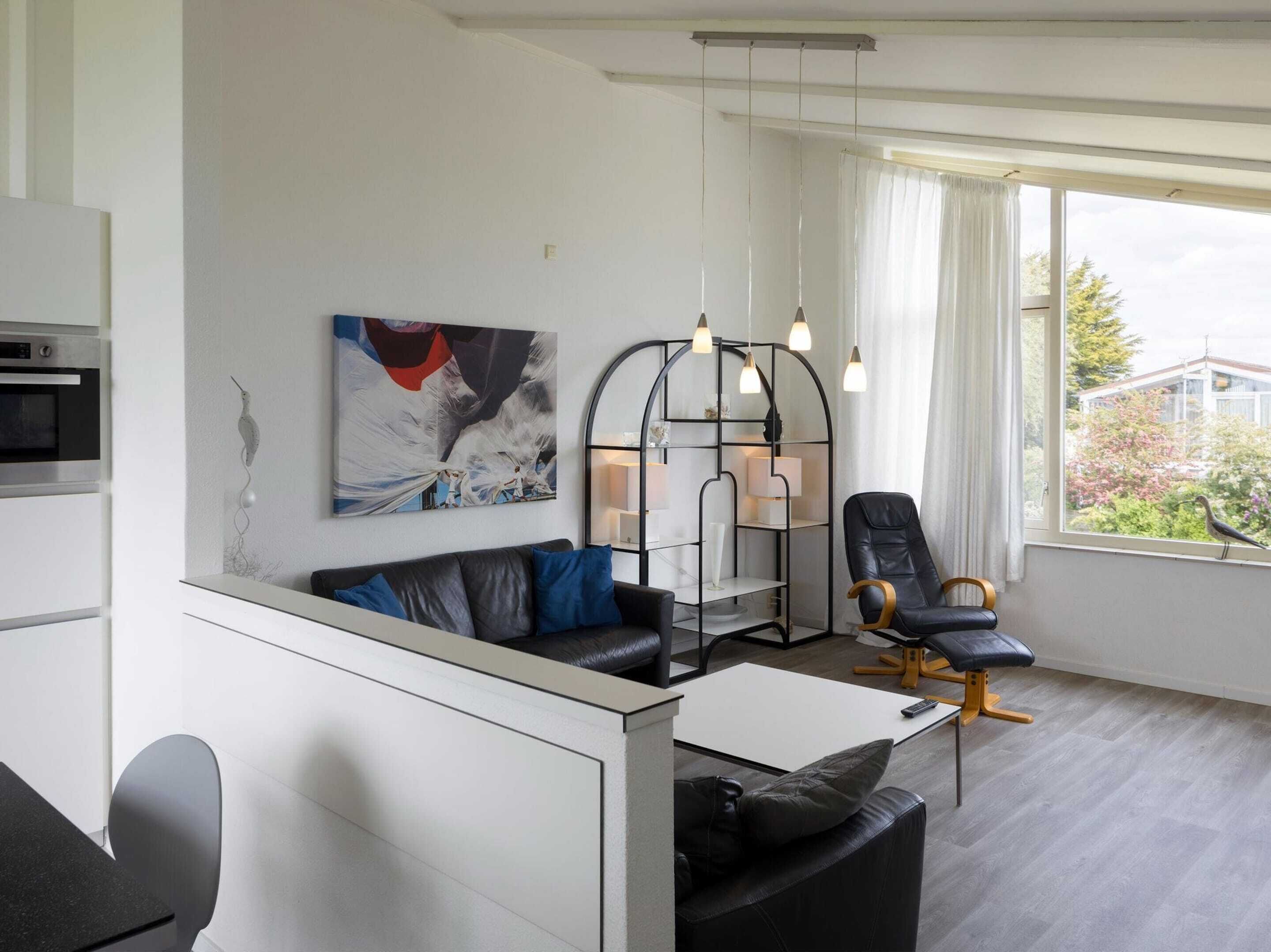 A modern living room with a black leather sofa, a white coffee table, and a black chair with a gold base. There is a large abstract painting on the wall, a black metal shelving unit with lamps, and a window with a view of greenery outside.