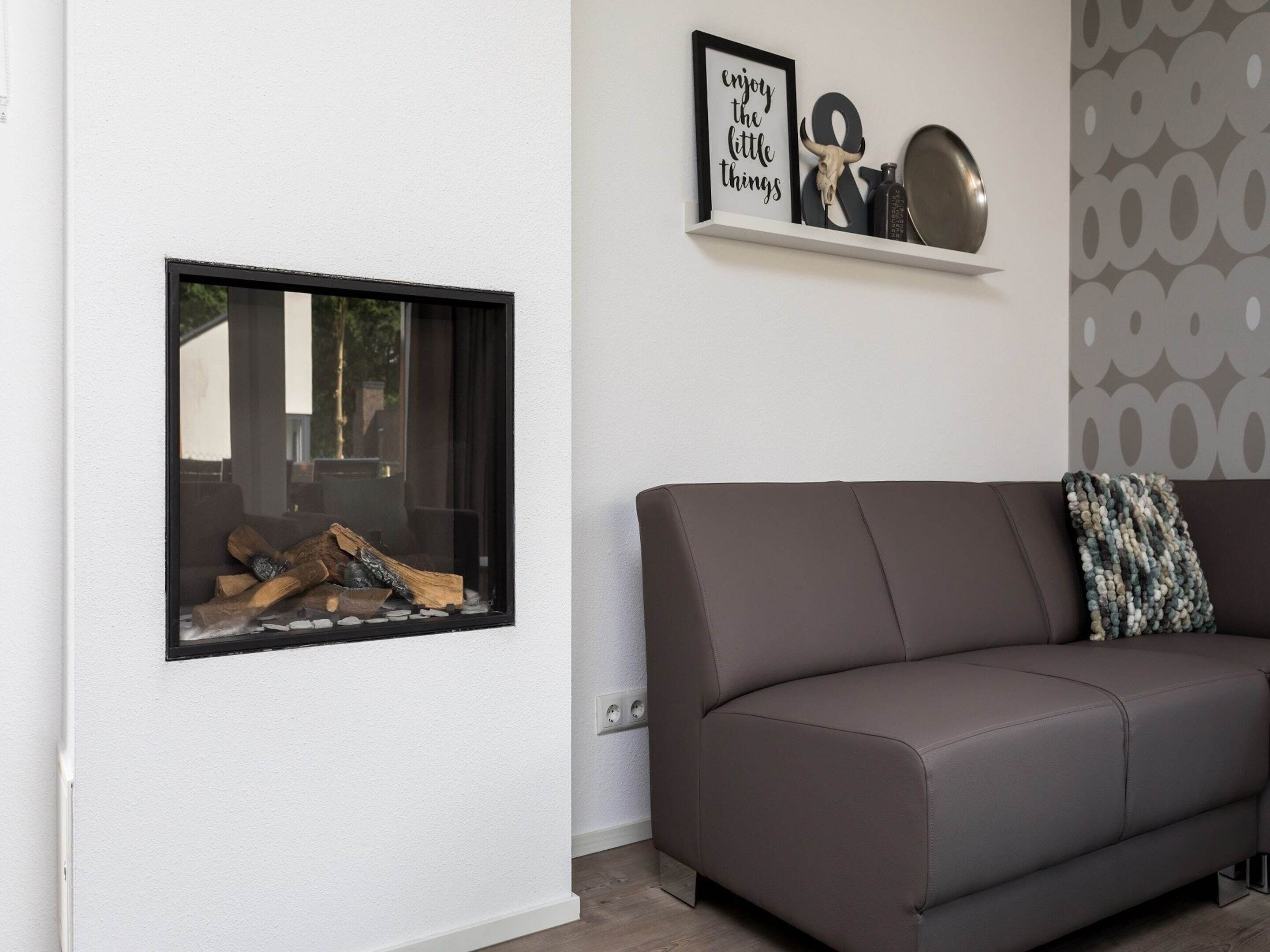 A modern living room with a brown leather sofa, a fireplace with logs, and a wall-mounted shelf with decorative items.