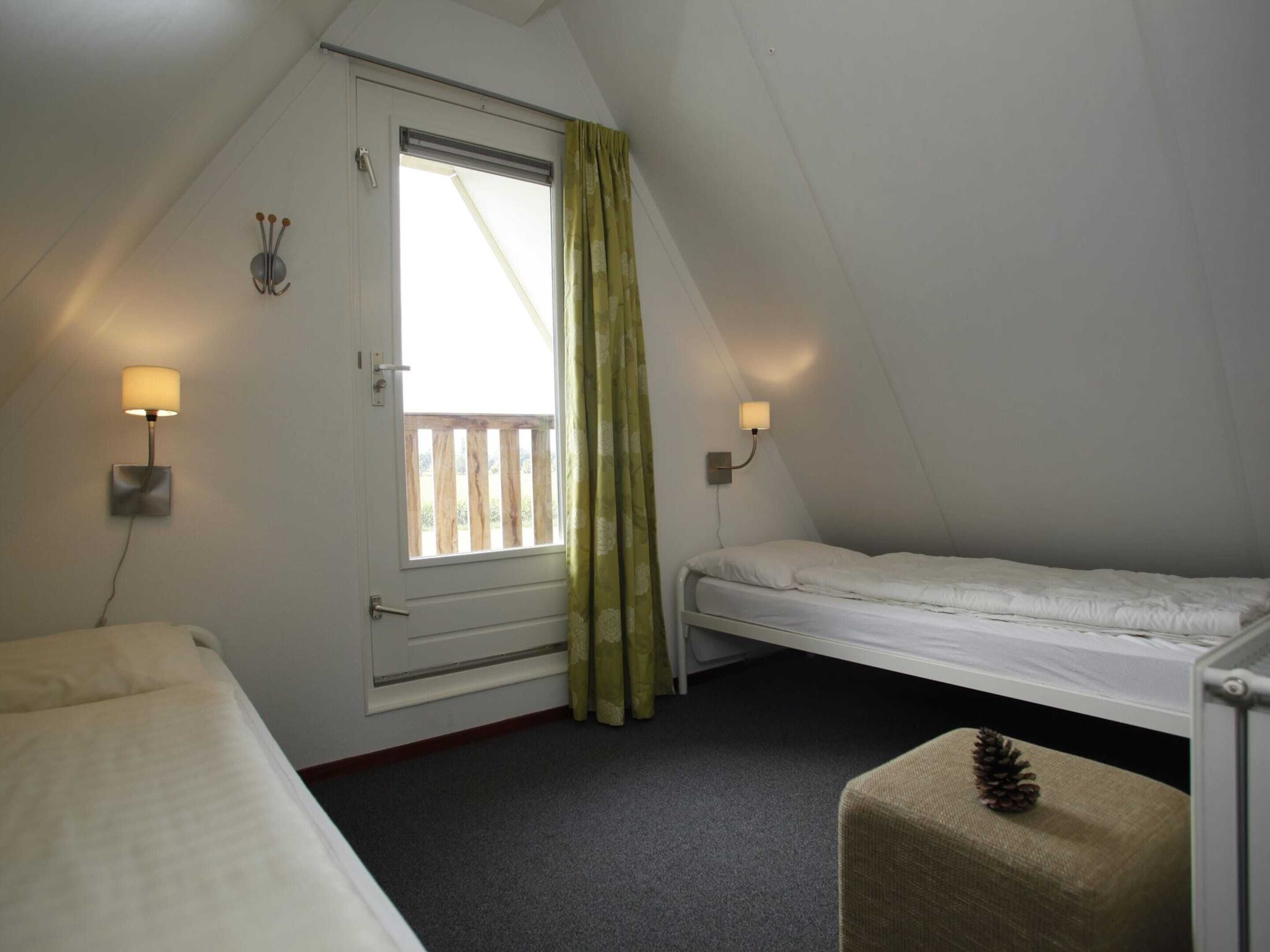 A small, triangular room with two beds, a window with curtains, and two wall-mounted lamps.