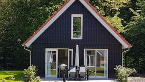 A dark-colored house with a red roof, large glass doors, and a small patio with outdoor furniture. - Modern Villa With Garden (Almen)