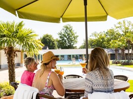 Un grupo de personas sentadas en una mesa al aire libre bajo un paraguas amarillo, con una piscina y follaje en el fondo.