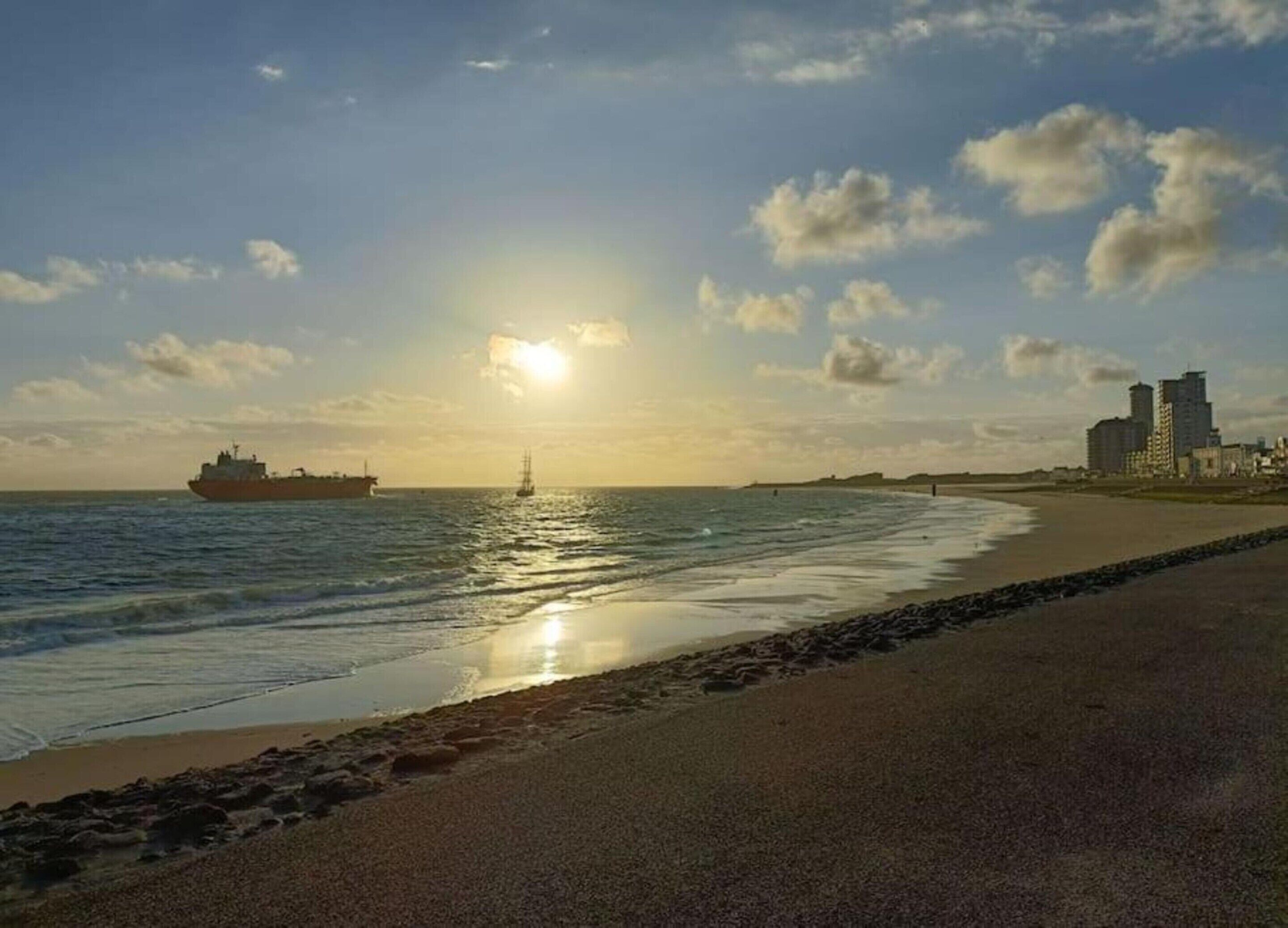 Een strand met een schip in het water, een heldere lucht en gebouwen op de achtergrond.