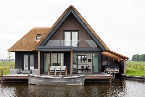 A modern house with a boat docked in front, large windows, and a thatched roof. - Luxury Villa on the Water (Ossenzijl)