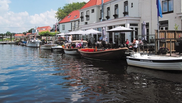 A waterfront area with boats docked along the shore, buildings with red roofs, and outdoor seating areas with umbrellas.