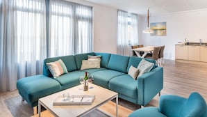Een moderne woonkamer met een blauwgroene modulaire bank, een houten salontafel en een eethoek met een witte tafel en stoelen.