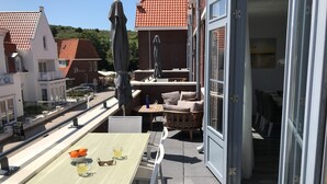 Een terras buiten met een tafel voor twee personen, een zithoek met stoelen, en een uitzicht op de straat en gebouwen op de achtergrond.
