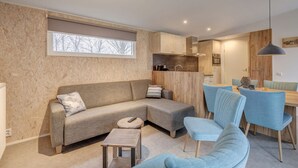 A modern living room with a sectional sofa, a small coffee table, and a dining area with blue chairs and a wooden table. - Modern Chalet With Dishwasher Near the Beach (The Hague)