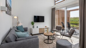 Un soggiorno moderno con un divano grigio, un tavolino da caffè e una TV a parete. Si vede una finestra con tende, una pianta e una vista sull'esterno.