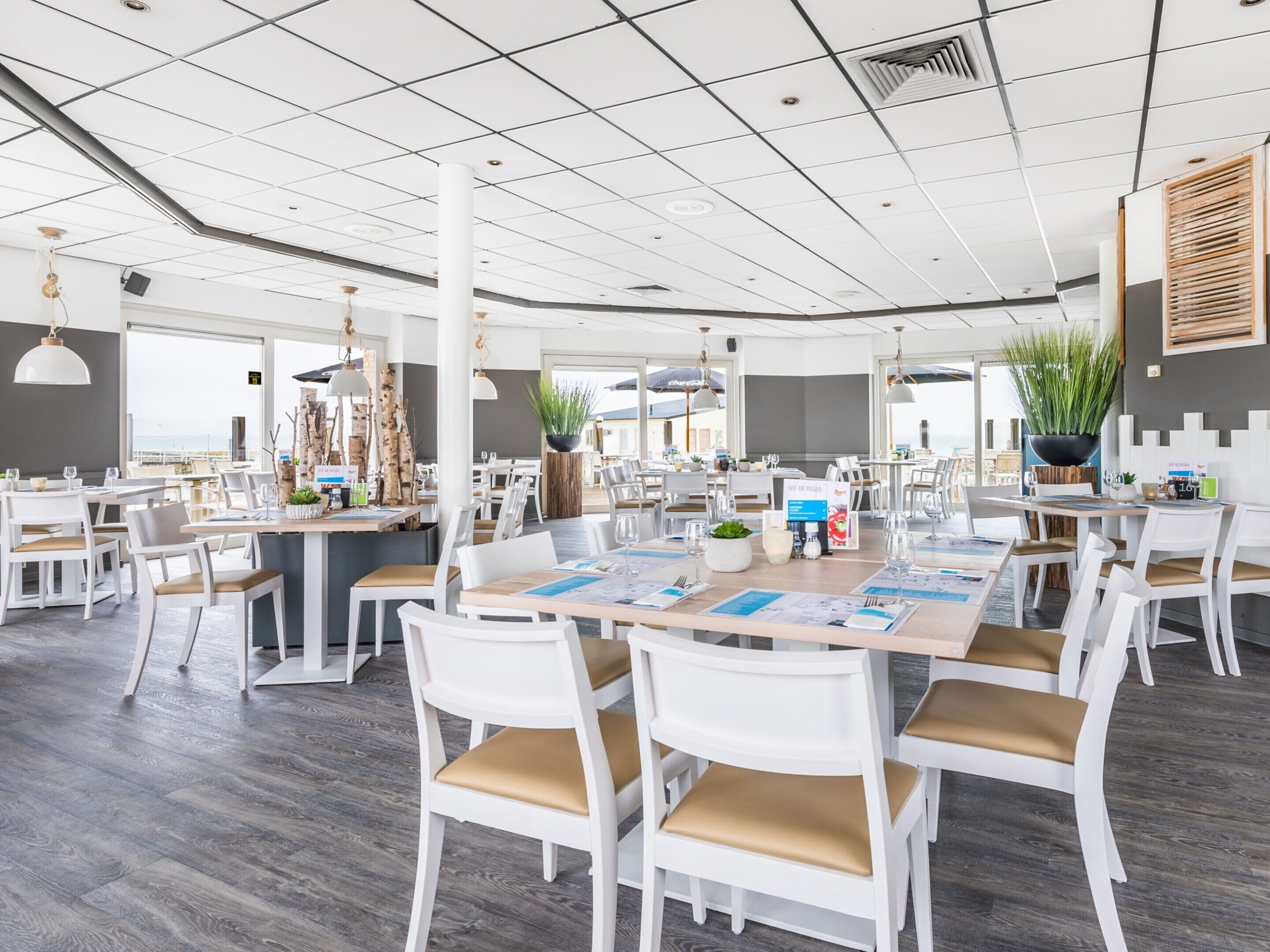 Un comedor amplio con mesas y sillas blancas, una isla central con una planta, y vista al mar a través de grandes ventanales.