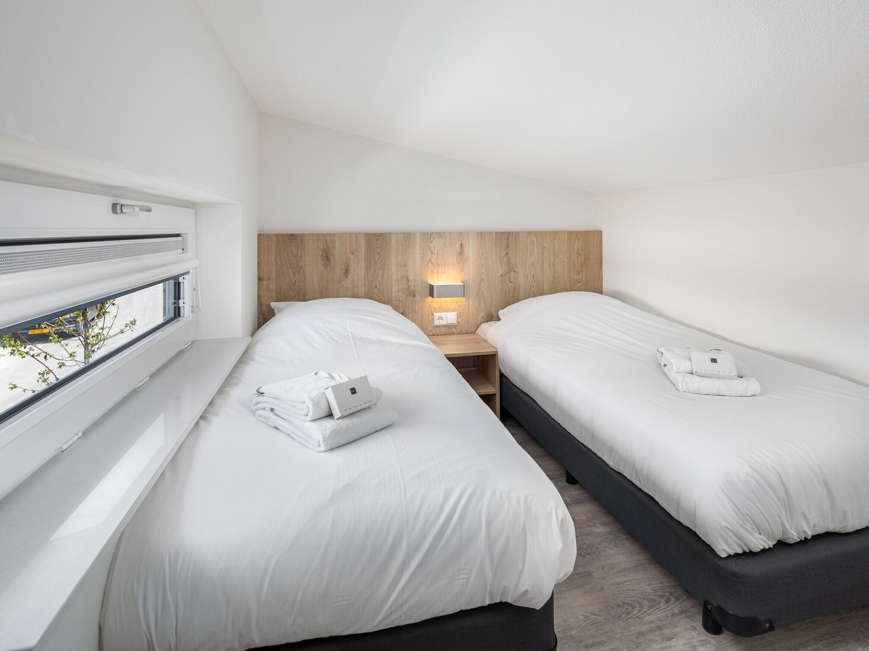 A modern hotel room with two beds, a wooden headboard, a bedside table with a lamp, and a window with a view of greenery.