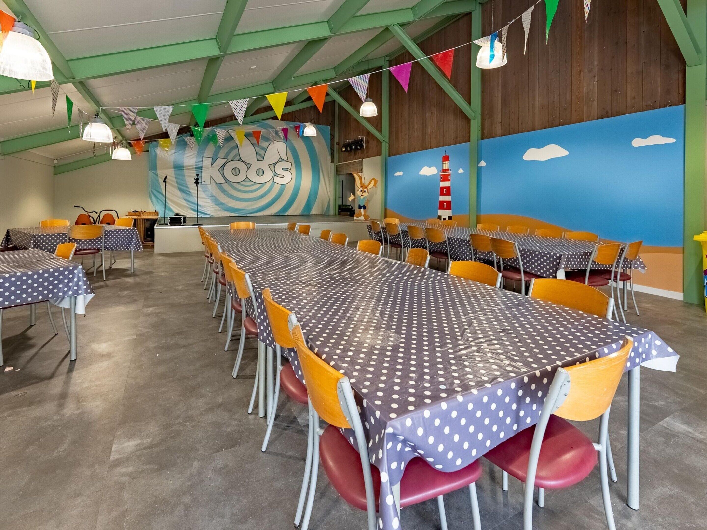 A spacious indoor dining area with multiple tables and chairs, featuring a colorful mural with a lighthouse and a rabbit, and a green and blue wall.