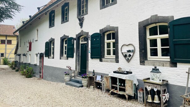 Een wit gebouw met groene luiken, een hartvormige decoratie en een klein terras met een tafel en stoelen.