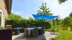 Een terras buiten met een tafel, stoelen, een blauwe parasol en een barbecue.