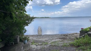 A gravel path leading to a wooden dock extending into a calm lake, surrounded by greenery and a clear sky. - Blue Spruce Lakefront Retreat - Recharge, reset, renew. (Alburgh)