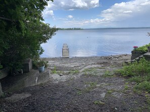 A gravel path leading to a wooden dock extending into a calm lake, surrounded by greenery and a clear sky.