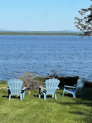 Three Adirondack chairs facing a lake with a forested shoreline and distant mountains.