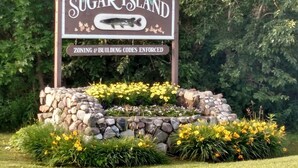 Sugar Island -saarelle johtava tervetulotoivotuskyltti, jossa on kalan kuva. Kylttiä ympäröi kivimuuri ja sen edessä on keltaisia kukkia.