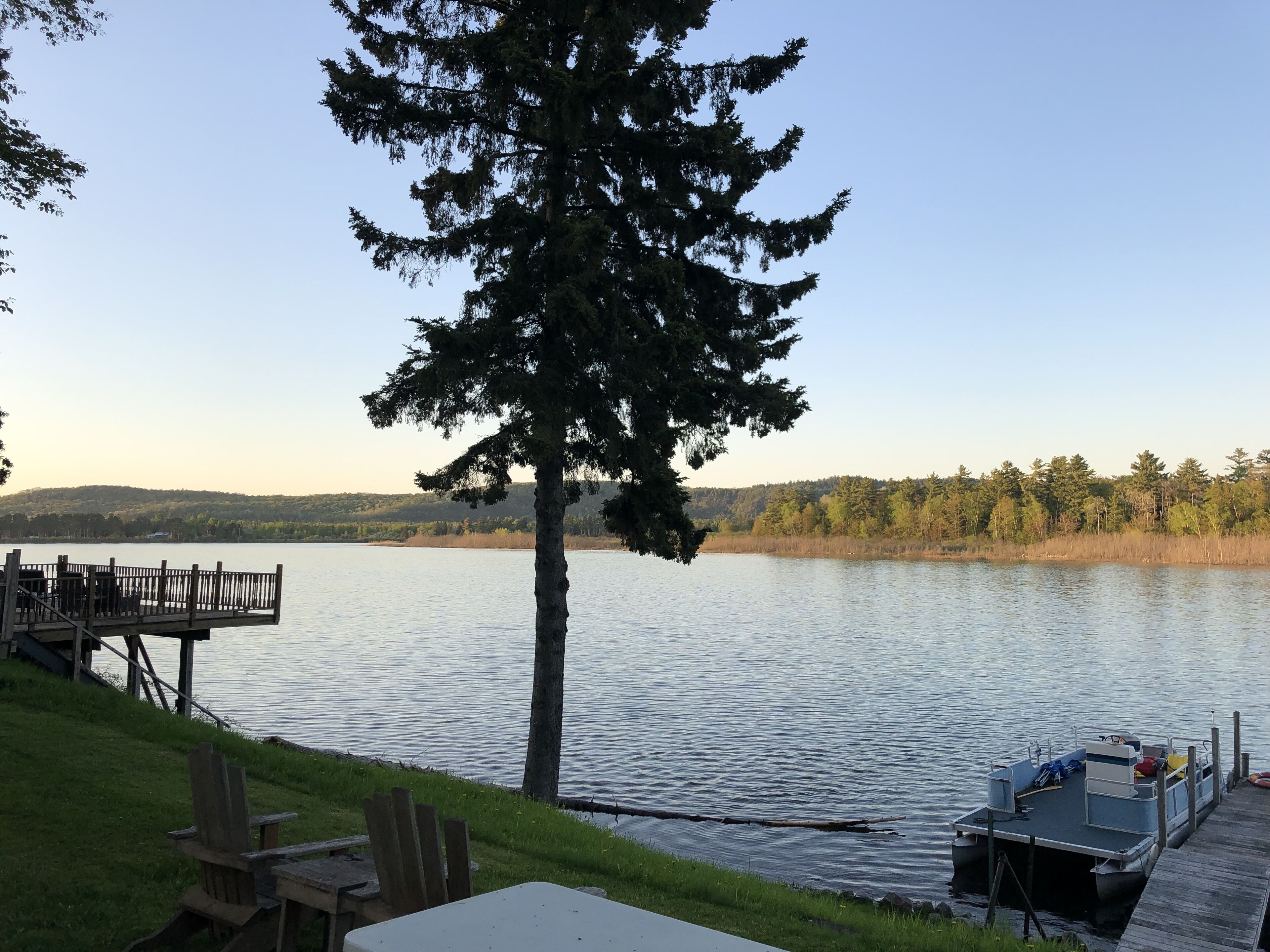 A lakeside deck with a table and chairs, a dock with a boat, and a large tree in the foreground.