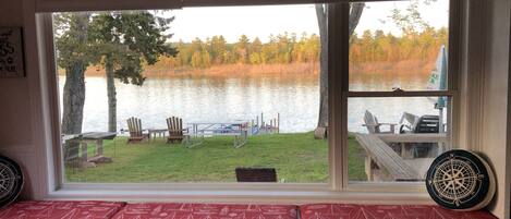 A room with a large window overlooking a lake, a wooden deck with chairs, and a bench with red cushions.