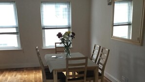 Un coin repas avec une table et des chaises en bois, un vase de fleurs et de grandes fenêtres équipées de stores.